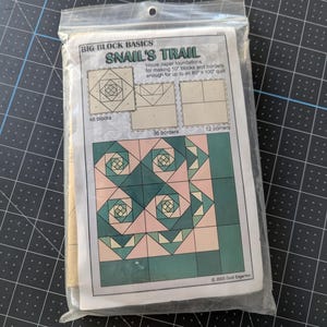 May include: A package of "Snail's Trail" quilt block tissue paper foundations. The design features a geometric pattern in shades of pink, green, and white. The package includes instructions for making 10" blocks and borders for an 80" x 100" quilt.