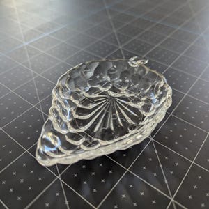 May include: Clear glass dish in the shape of a pinecone. The dish has a textured surface with a detailed pattern. The dish is sitting on a dark surface.