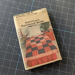 May include: A vintage sewing pattern titled "Americana Patchwork Quilts" by Butterick. The pattern features a quilt design with red, white, and blue squares and stripes. The pattern is in a rectangular package.