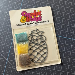 May include: A Makit & Bakit stained glass suncatcher craft kit featuring a pineapple-shaped metal frame and three small plastic bags containing green, yellow, and brown translucent crystals for melting, all packaged on a branded cardboard backing.