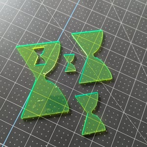 May include: A collection of neon green acrylic hourglass shapes in various sizes. The shapes are arranged on a black cutting mat with a grid pattern. The hourglasses are transparent and have a geometric design.