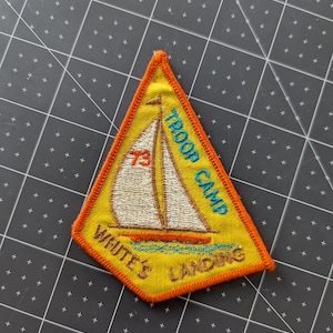 May include: A yellow triangular patch with an orange border. The patch features a sailboat with the text "TROOP CAMP" and "WHITE'S LANDING". The number "73" is visible on the sail. The patch is likely for a scouting event.