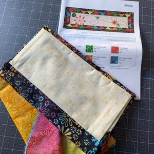 May include: A folded table runner with a floral design, featuring a cream-colored center with a floral pattern, bordered by black fabric with colorful flower prints. A pattern sheet labeled "Flower Power Table Runner" is visible.