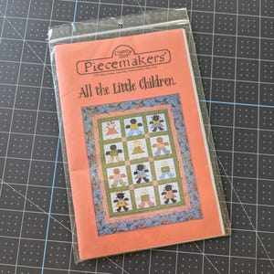 May include: A Country Store Piecemakers pattern booklet titled All the Little Children, featuring a quilt design with twelve stylized figures in a grid, enclosed in a clear plastic sleeve with a peach-colored cover and floral border.