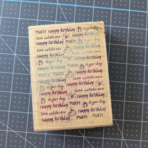 May include: A rectangular wooden rubber stamp featuring a repeating pattern of birthday-themed text and icons. The text includes phrases like Happy Birthday, Party, Let's Celebrate, and It's Your Day, interspersed with small illustrations of gifts and cakes.