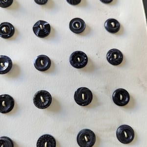 May include: A collection of twenty-one black circular buttons arranged in a grid pattern on a light-colored backing. Each button features a distinct textured or patterned surface and a central hole for attachment, showcasing various rim designs.
