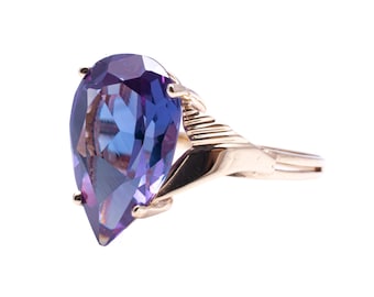 May include: A gold ring featuring a large, pear-shaped gemstone with shades of purple and blue. The ring's band has a textured design near the stone, and the setting is gold-colored.