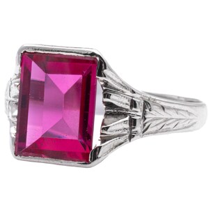 May include: A silver ring featuring a large, rectangular, bright pink gemstone. The ring has an ornate design with etched details on the band and around the gemstone setting. The gemstone is a vibrant, eye-catching color.