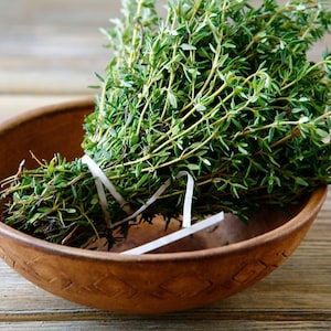 May include: A bundle of fresh thyme, tied with a white ribbon, rests in a rustic, wooden bowl. The thyme has small, green leaves and thin, light brown stems. The bowl is a warm, earthy brown color, and the background is a wooden surface.