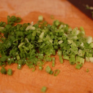 May include: A close-up of a wooden cutting board with finely chopped green onions. The onions are arranged in a pile on the board.