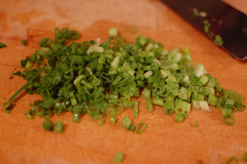 May include: A close-up of a wooden cutting board with finely chopped green onions. The onions are arranged in a pile on the board.