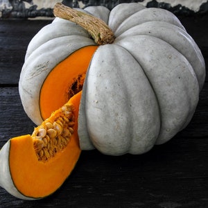 May include: A gray pumpkin with a light orange interior is cut in half and a slice is removed. The pumpkin is sitting on a black surface.