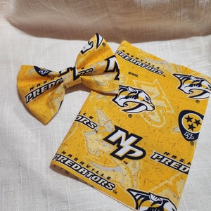 May include: A yellow bow tie with a black and white Nashville Predators logo. The bow tie is made of fabric and has a textured surface. The bow tie is paired with a yellow fabric square with the Nashville Predators logo.