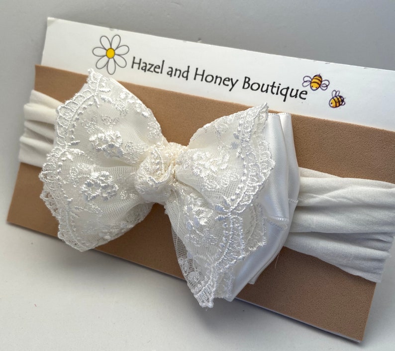 Puede incluir: Diadema de encaje blanco con una banda el&aacute;stica blanca. La diadema est&aacute; sobre una tarjeta marr&oacute;n con el texto "Hazel and Honey Boutique" y una margarita.