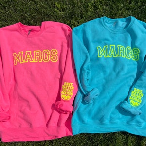 May include: Two long-sleeved sweatshirts, one pink and one blue, with the word "MARGS" printed in yellow on the front. The sleeves have the text "SAVE WATER DRINK MARGS" and a lemon graphic printed in yellow.