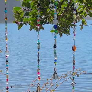 May include: Four vertical strands of multi-colored glass and plastic beads hanging from a tree branch. Each strand features a variety of shapes, sizes, and colors, including clear, blue, green, red, and amber beads, with small metal bells at the ends.