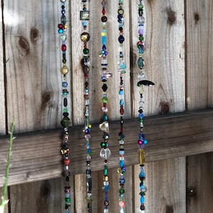 May include: A handmade wind chime with multiple strands of colorful beads and small bells. The beads are various shapes and colors, including blue, green, gold, and clear. The chime hangs from a natural wooden branch.