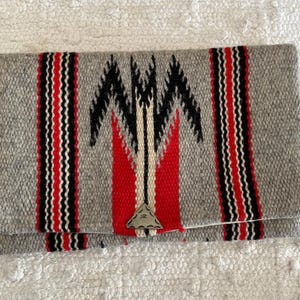 May include: A gray woven clutch with a geometric design in black, red, and white. The clutch features vertical stripes and a triangular metal clasp. The folded design is rectangular and rests on a white textured surface.