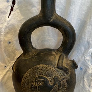 Puede incluir: Un recipiente de cerámica marrón oscuro con base redondeada, cuello largo y asa. El recipiente presenta un diseño circular con una figura de ave. La pieza tiene una forma única y es probablemente un objeto decorativo.