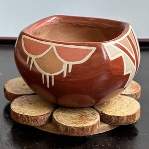 May include: A small, rounded terracotta-colored clay bowl featuring hand-painted geometric and scalloped white and tan patterns around the exterior rim. The bowl is resting on a circular base constructed from several small, flat, natural wood slices.