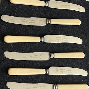 May include: A set of six vintage knives with cream-colored handles and stainless steel blades. The blades are marked with text, and the knives are arranged on a dark surface. The knives are likely for a formal dining setting.