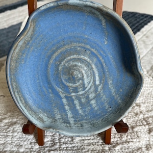 Puede incluir: Un reposacucharas de cerámica azul hecho a mano con una forma orgánica. El reposacucharas presenta un diseño en espiral en el centro y un borde azul claro. La pieza de cerámica se exhibe sobre un pequeño soporte de madera.