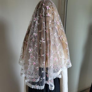 May include: A sheer white veil, adorned with pink floral embroidery and a scalloped lace edge. The veil is draped over the head and shoulders, creating a soft, romantic effect. Suitable for a wedding or special event, the veil is a delicate accessory.