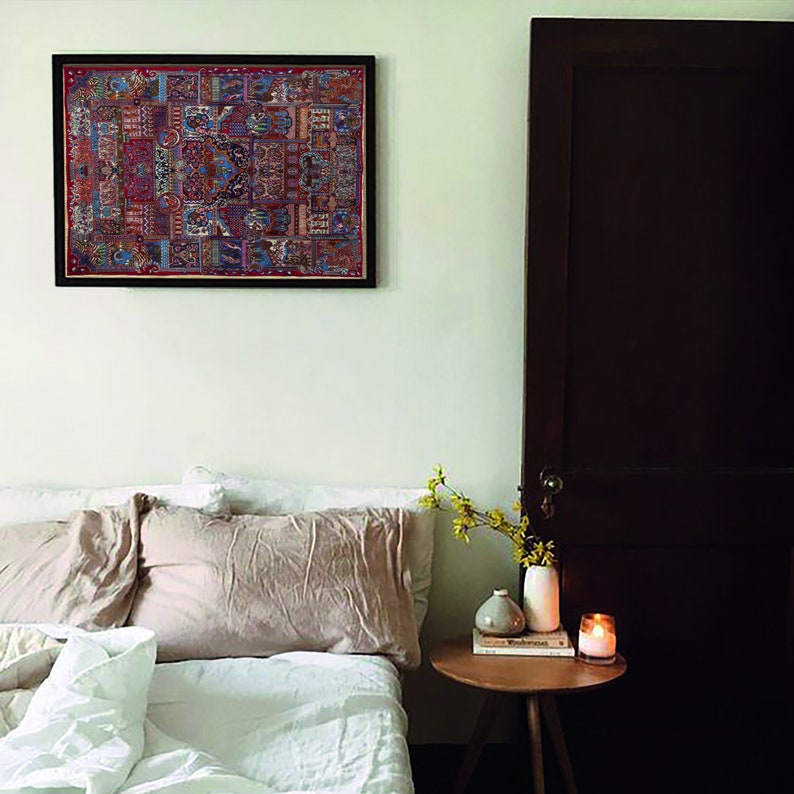 May include: A white bedroom with a bed, a nightstand, and a framed tapestry hanging on the wall. The bed has a white duvet and two pillows, one beige and one white. The nightstand is made of wood and has a vase of yellow flowers, a candle, and a book on it. The tapestry is a colorful, intricate design with a red, blue, and gold color scheme.