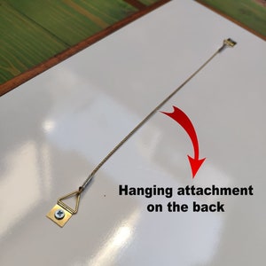 May include: A close-up of a white surface with a gold-colored hanging attachment and a string attached to it. The text "Hanging attachment on the back" is written in black below the attachment.