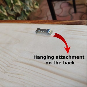May include: Close-up of a wooden sign with a metal hanging attachment on the back. The text "Hanging attachment on the back" is printed on the wood.
