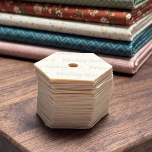 May include: A stack of hexagonal paper templates with the text "Prickly Notions" and a 1-inch hole, sits in front of a stack of colorful fabric. The fabric includes green, orange floral, cream with gold flecks, teal, and pink.