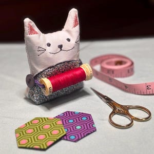 May include: A handmade cat-shaped pin cushion with a red spool of thread, a pink measuring tape, and small gold scissors. Two hexagon-shaped fabric pieces are also present. The cat pin cushion has a floral patterned base.