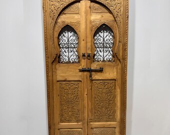 May include: A wooden double door with intricate carvings and an arched top. The door features decorative ironwork behind the arched openings and a black metal latch. The wood has a natural, light brown finish.