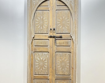 May include: A pair of intricately carved wooden doors with a whitewashed finish. The doors feature a traditional Moroccan design with geometric patterns and an arched top.