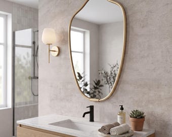 May include: An irregular, organic-shaped wall mirror with a thin gold frame. The mirror is mounted above a wooden vanity cabinet featuring a white marble countertop, a black faucet, a soap dispenser, two rolled towels, and a small potted succulent.
