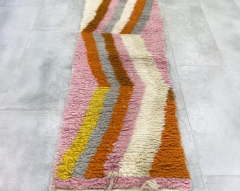May include: A colorful, textured runner rug with a zig-zag pattern. The rug features stripes of pink, yellow, orange, gray, brown, and white. The rug has a fringe at the bottom. This rug is suitable for a hallway or entryway.
