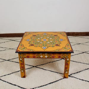 Peut inclure: Une table basse carrée de couleur jaune avec des motifs floraux peints à la main complexes dans des tons de vert, bleu, rouge et blanc. La table a quatre pieds et un plateau brun foncé.