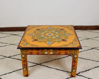 May include: A square, yellow coffee table with intricate hand-painted floral designs in shades of green, blue, red, and white. The table has four legs and a dark brown top.