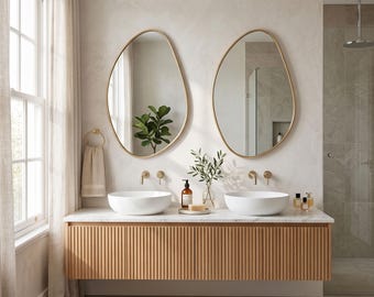 May include: A bathroom vanity featuring a light wood cabinet with vertical slats, a white marble countertop, and two white vessel sinks. Two asymmetrical mirrors with thin gold frames are mounted above the sinks with gold wall-mounted faucets.