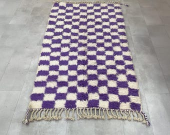 May include: A rectangular, hand-woven rug with a checkerboard pattern of purple and cream squares. The rug has a shaggy texture and a fringe along the short ends. The rug is approximately 36 inches wide and 60 inches long.