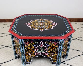May include: Octagonal wooden coffee table with intricate hand-painted floral designs in shades of gold, blue, pink, and green on a black background. The table has a red border and a carved base.