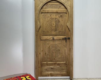 May include: A carved wooden door with intricate designs. The door is made of light brown wood and has a dark brown metal handle. The door is set against a white wall.