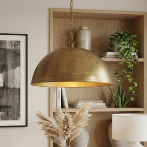 May include: A large, gold-colored pendant light with a dome-shaped shade hangs from a chain. The light fixture is suspended in front of a wooden bookshelf filled with books and plants. A framed black and white artwork is on the left.