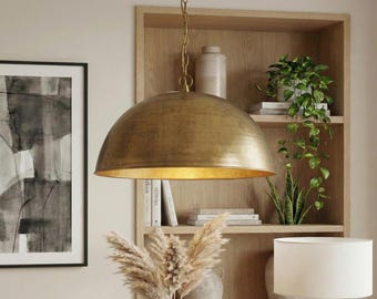 May include: A large, gold-colored pendant light with a dome-shaped shade hangs from a chain. The light fixture is suspended in front of a wooden bookshelf filled with books and plants. A framed black and white artwork is on the left.