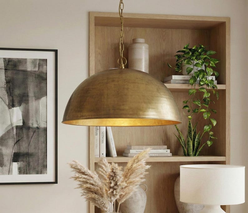 May include: A large, gold-colored pendant light with a dome-shaped shade hangs from a chain. The light fixture is suspended in front of a wooden bookshelf filled with books and plants. A framed black and white artwork is on the left.