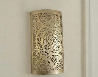 May include: A gold-colored wall sconce with a cylindrical shape. The sconce features an intricate, openwork design of geometric shapes, including circles and triangles. It is mounted on a white wall, adding a decorative element to the space.