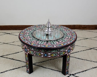 May include: A round, black wooden table with intricate painted designs in shades of pink, green, and blue. The table has four legs and a silver tray with a teapot and glasses on top.