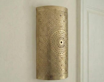 May include: A brass wall sconce with a cylindrical shape and a decorative, perforated design. The sconce features a central circular pattern and is mounted on a white wall. The fixture is designed to hold a light source.