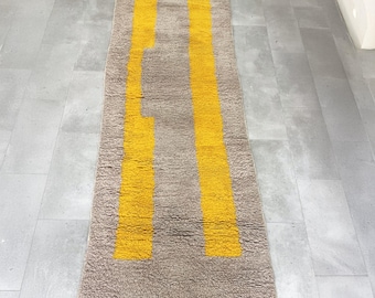 May include: A rectangular, beige rug with two vertical, bright yellow stripes. The rug has a textured surface and a fringe along the bottom edge. The design is modern and minimalist, suitable for a hallway or entryway.