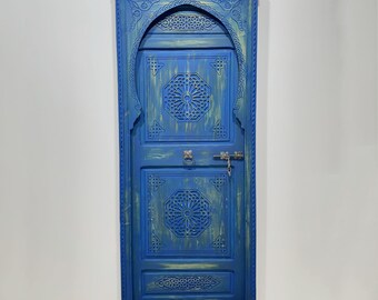 May include: A blue wooden door with intricate carvings and a brass handle. The door has a distressed finish and is framed by a decorative archway.